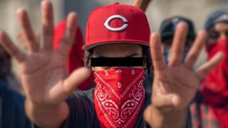 Red bandana-wearing protester with raised hands and a baseball cap, face partially covered.